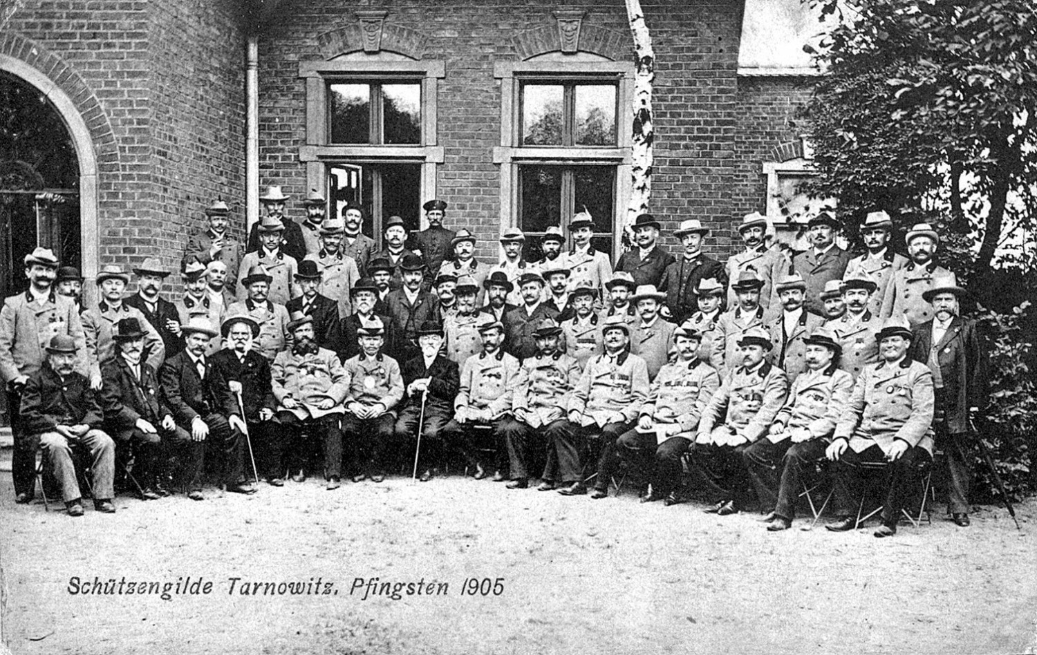 Członkowie tarnogórskiego bractwa przed budynkiem strzelnicy. Pocztówka pamiątkowa z zawodów w Zielone Świątki 1905 r. Źródło: Historia Tarnowskich Gór, red. J. Drabina, Tarnowskie Góry 2000. Członkowie tarnogórskiego bractwa przed budynkiem strzelnicy. Pocztówka pamiątkowa z zawodów w Zielone Świątki 1905 r. Źródło: Historia Tarnowskich Gór, red. J. Drabina, Tarnowskie Góry 2000.